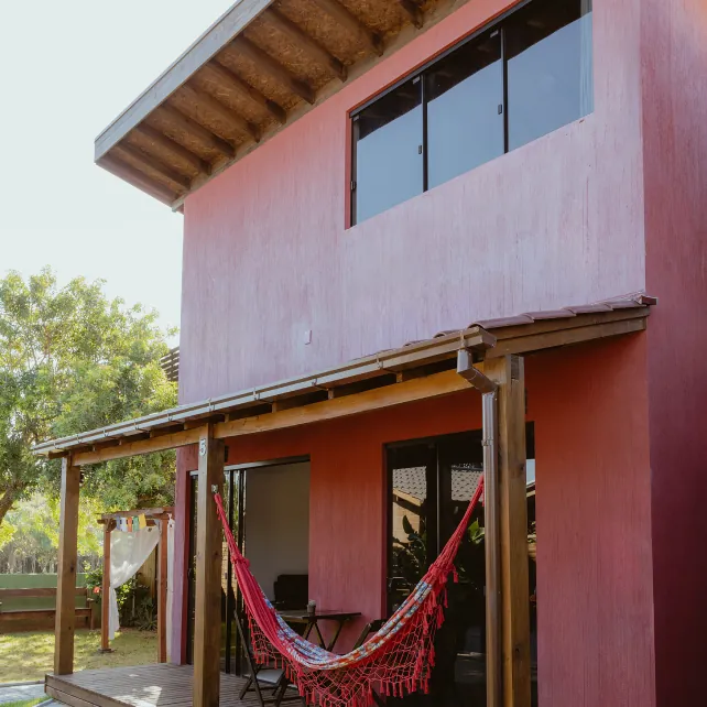 Fachada rosa da casa, com varanda de madeira e rede vermelha armada.
