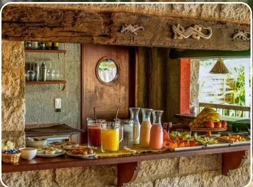 Mesa de café da manhã da pousada com sucos, frutas e pães sobre bancada de pedra.