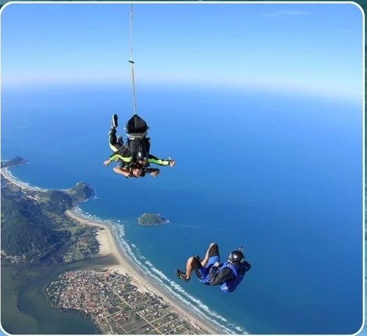 Salto de paraquedas duplo sobre a costa da Praia do Rosa, com a lagoa e o mar ao fundo.