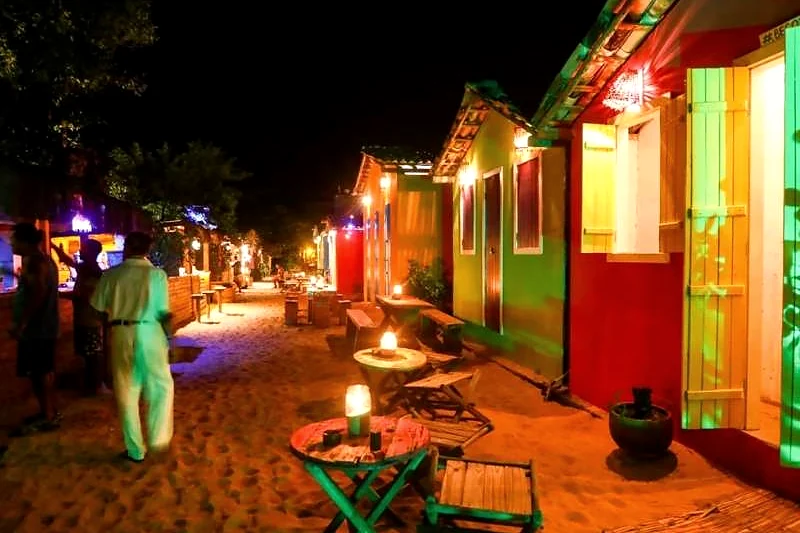 Ruas de areia de Caraíva à noite, com casas coloridas iluminadas e mesas na rua.