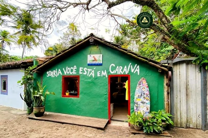 Casa verde de madeira na vila com a frase pintada: aqui você está em Caraíva.