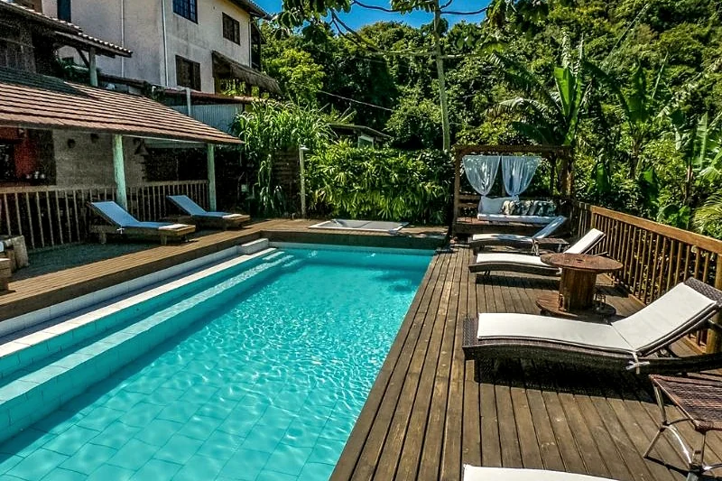 Piscina com deck de madeira e espreguiçadeiras cercada por vegetação.