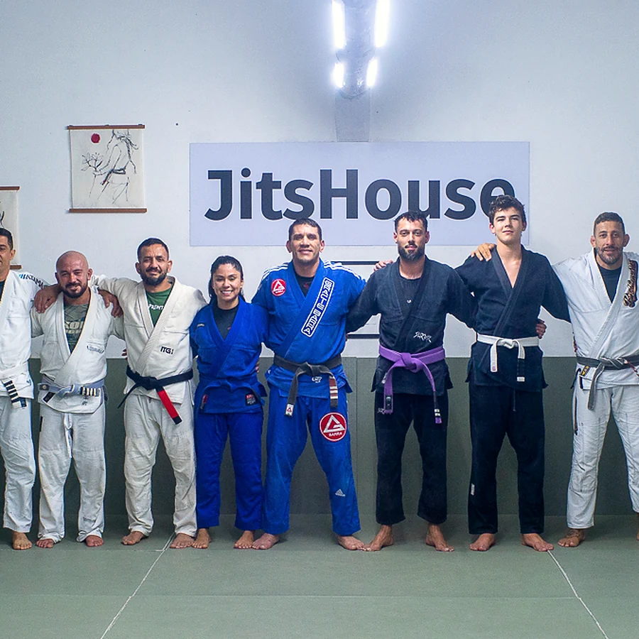 Turma enfileirada de kimono no tatame, diante do selo e do painel da JitsHouse.