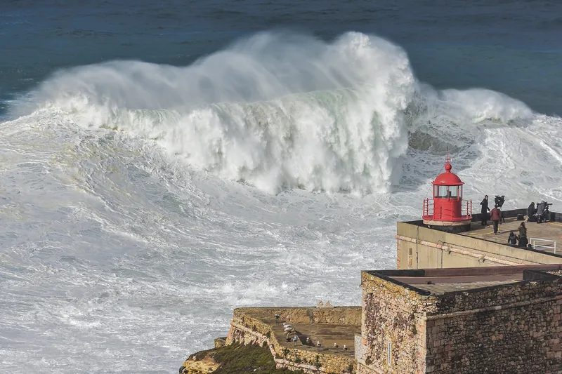 O farol vermelho de Nazaré sobre o forte de pedra, com uma onda gigante estourando atrás.
