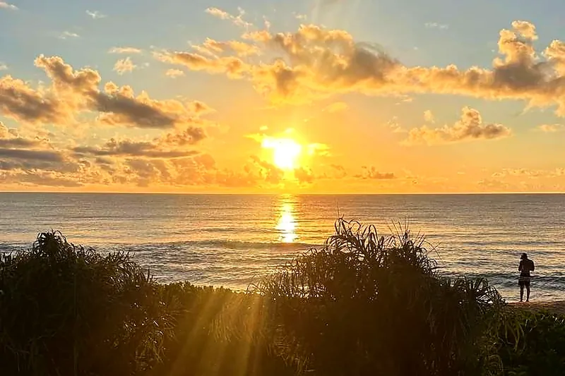 Sol baixo sobre o mar na Praia do Rosa, com a restinga em silhueta e uma pessoa sozinha na areia.