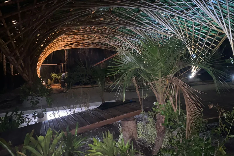 Detalhe dos arcos de bambu à noite, com o jardim e a passarela de madeira em primeiro plano.