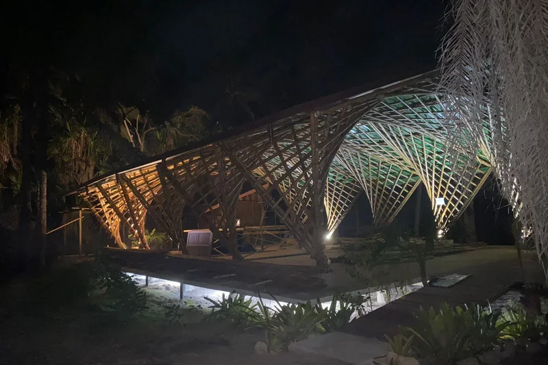 A estrutura de bambu vista de fora à noite, iluminada por baixo, cercada de mata.