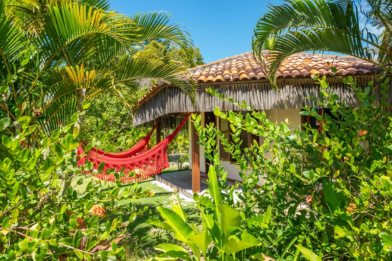 Bangalô de telhado de palha no meio do jardim, com uma rede vermelha armada na varanda.