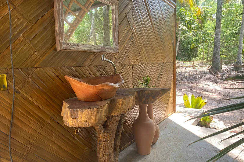 Lavabo dos vestiários: cuba de madeira sobre tronco, espelho e parede de bambu trançado.